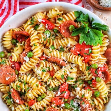 white bowl filled with pasta, tomatoes, herbs, peas, and linguica with red striped napkin..