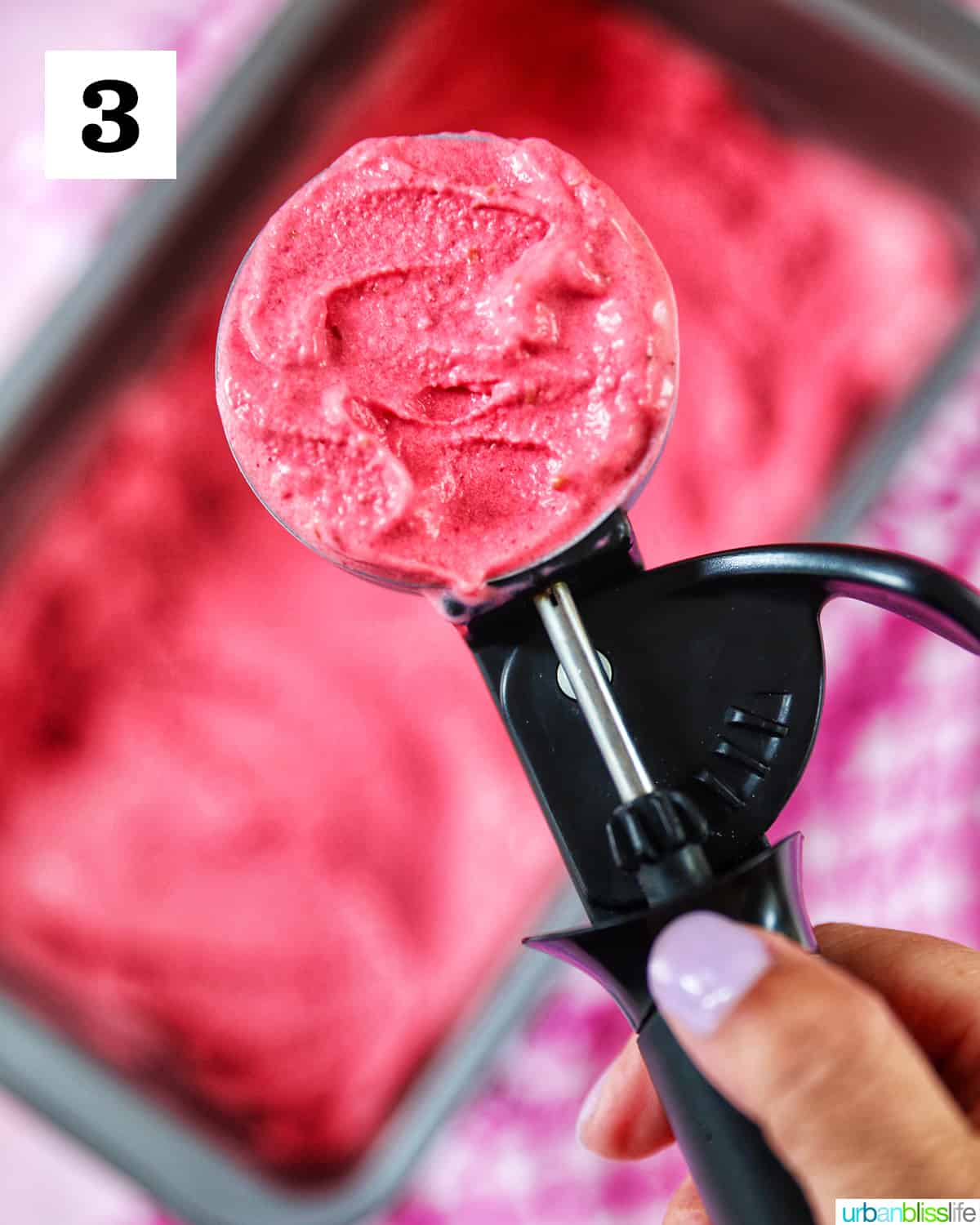 large ice cream scoop filled with raspberry sherbet over a pan filled with more raspberry sherbet blurred in the background.