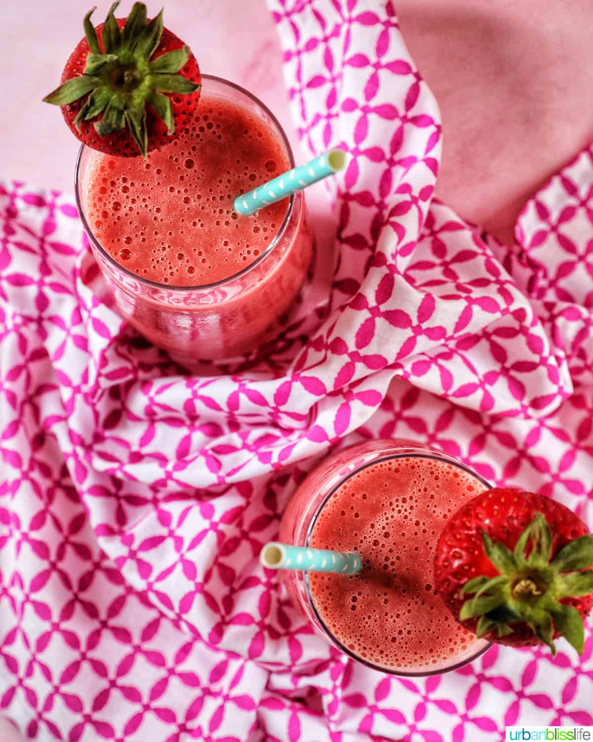 overhead photo of two glasses with strawberry watermelon smoothies, blue straws, sliced strawberry garnish on a pink and white napkin..