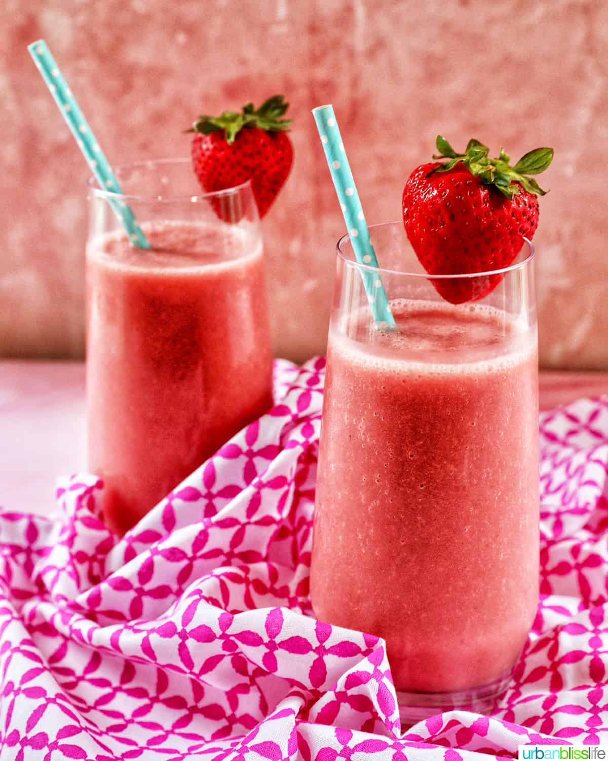 two glasses with strawberry watermelon smoothies, blue straws, sliced strawberry garnish on a pink and white napkin.