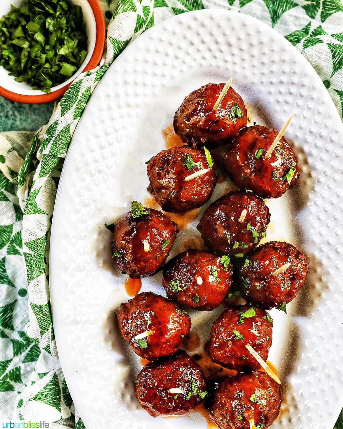 several honey bourbon meatballs with toothpicks on a white serving platter surrounded by a green and white napkin with small bowl of chopped parsley at the top left.