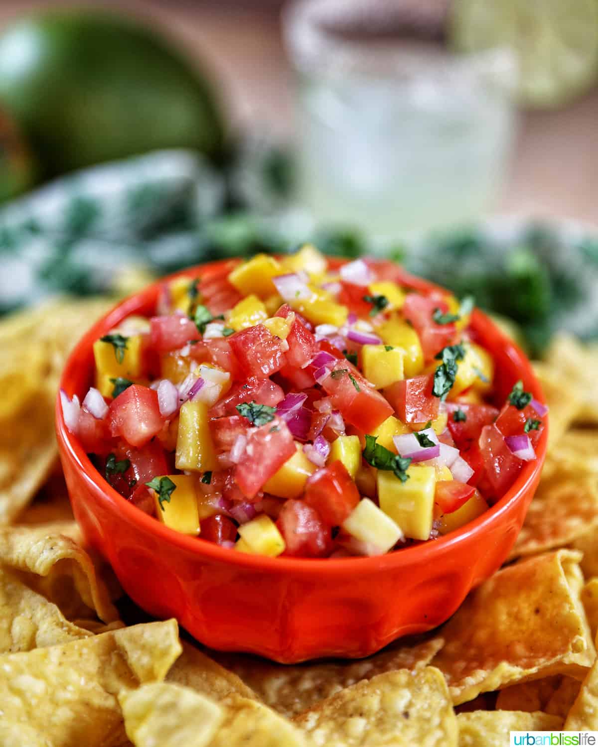 side angle of orange bowl filled with mango pico de gallo with mangos and margarita in the background.