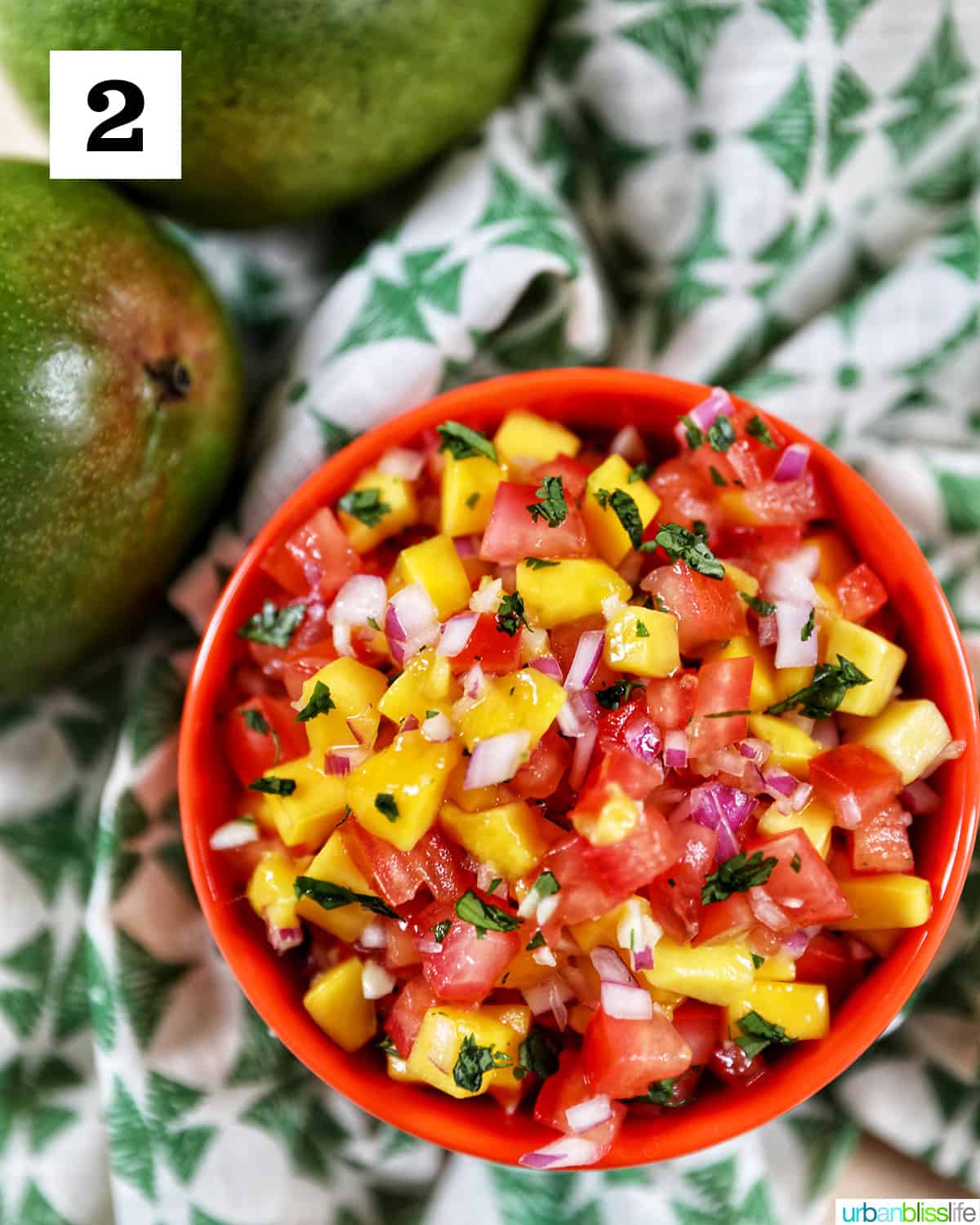 side angle of orange bowl filled with mango pico de gallo with mangos on the side on a green and white tablecloth.