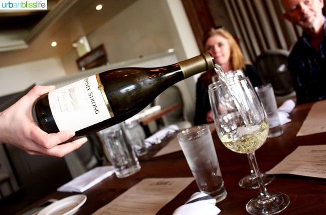 bottle of white wine pouring into a glass with people in the background.