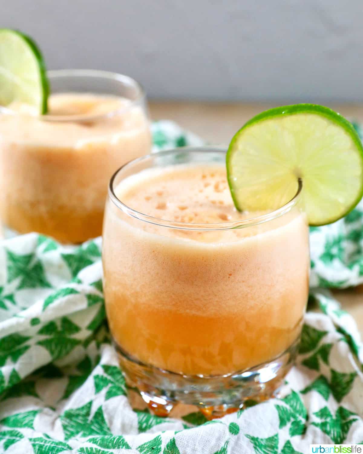 side view of two glasses of cantaloupe margaritas with lime slice garnish on a green and white patterned napkin.