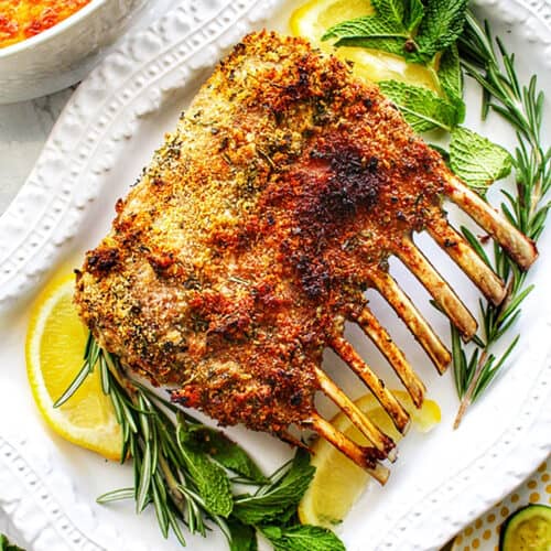 rack of lamb with lemon wedges and fresh herbs on a white platter.