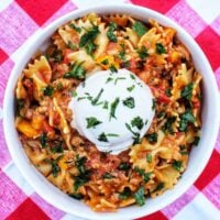 Taco Pasta with scoop of sour cream and chopped parsley on a red and white checkered tablecloth.