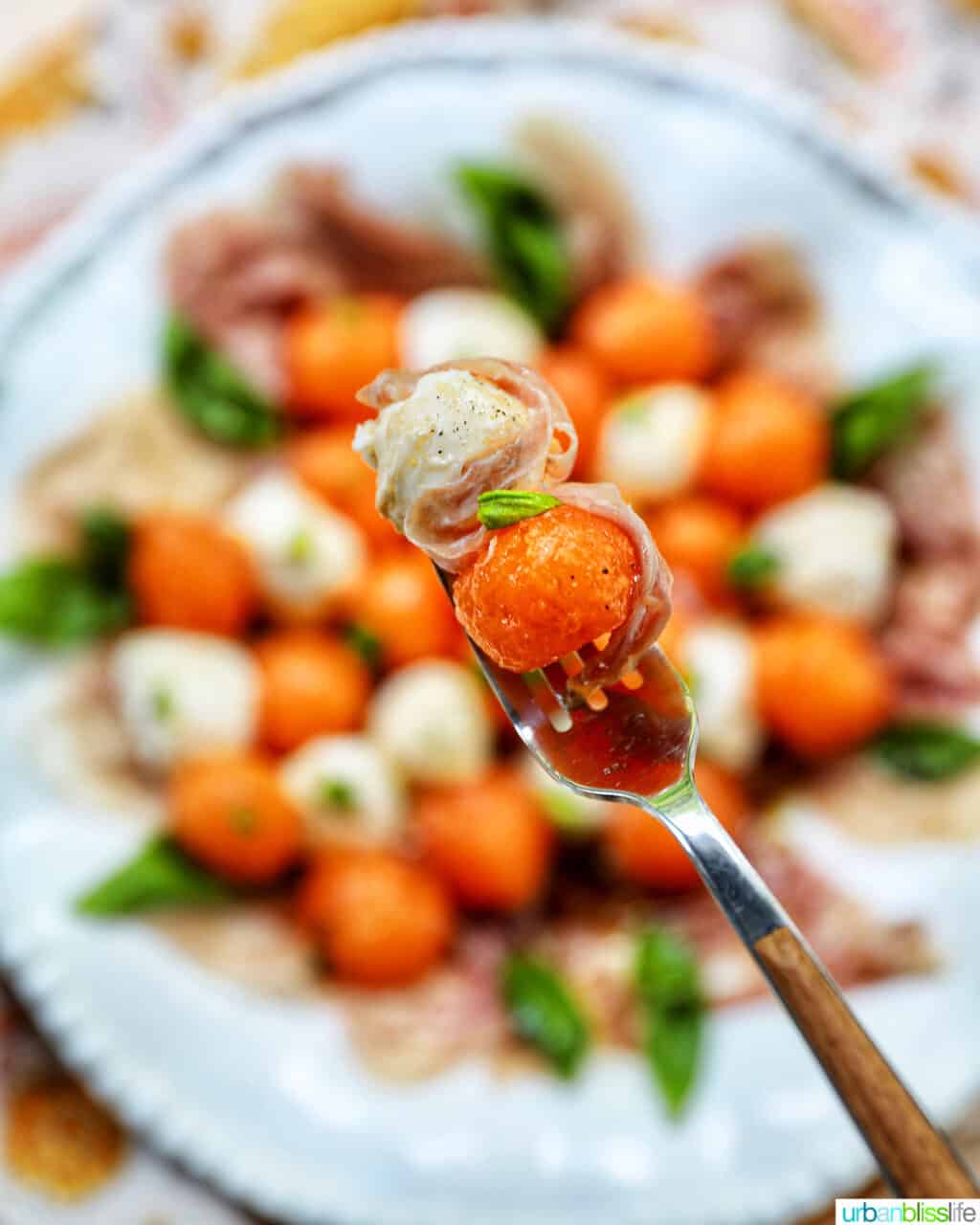 fork holding cantaloupe, mozzarella ball, prosciutto and basil over blurred bowl of melon prosciutto caprese salad.