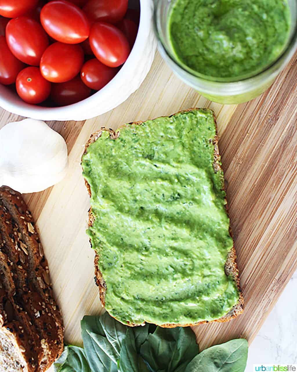 avocado pesto spread on a piece of bread, with jars of tomatoes, pesto, cheese, and spinach leaves places around the bread on a cutting board.