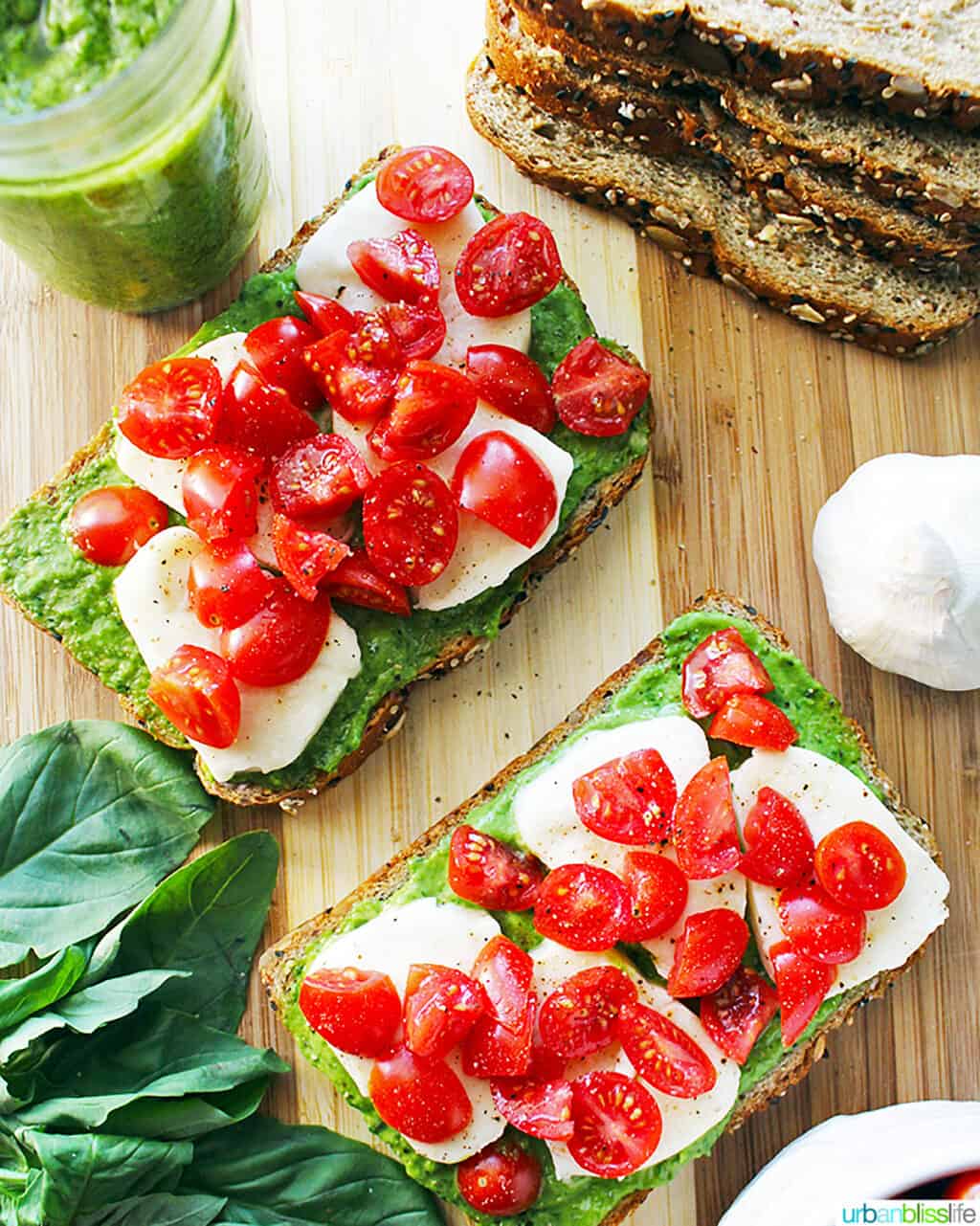 two pieces of pesto, cheese, tomato toast on a cutting board with slices of bread in one corner and spinach leaves in another corner.