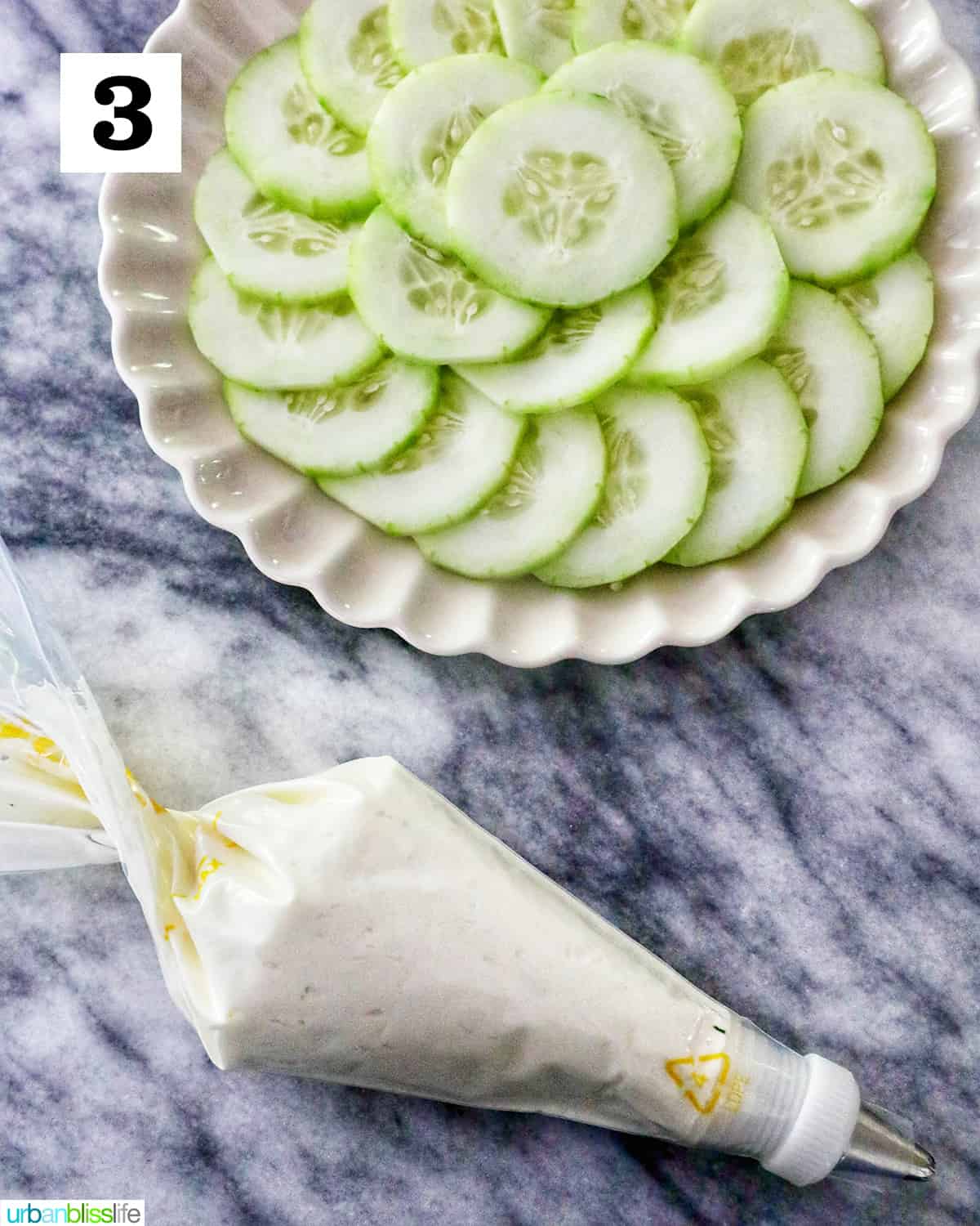 plate with fanned out cucumber slices next to a pastry bag with cream cheese on a marble countertop.