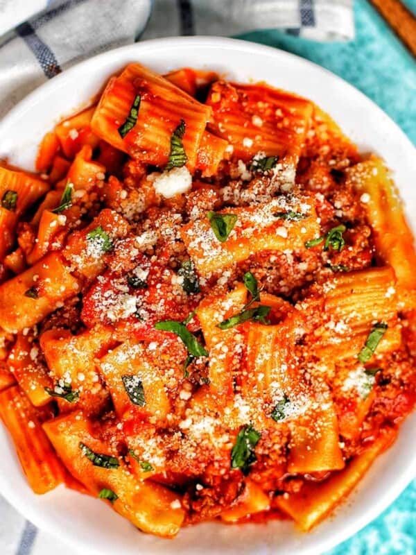 bowl of rigatoni pasta with Italian sausage on a white and blue checkered napkin and a fork.