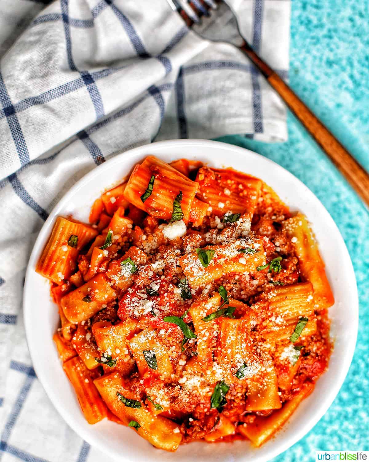 bowl of rigatoni pasta with Italian sausage on a white and blue checkered napkin and a fork.