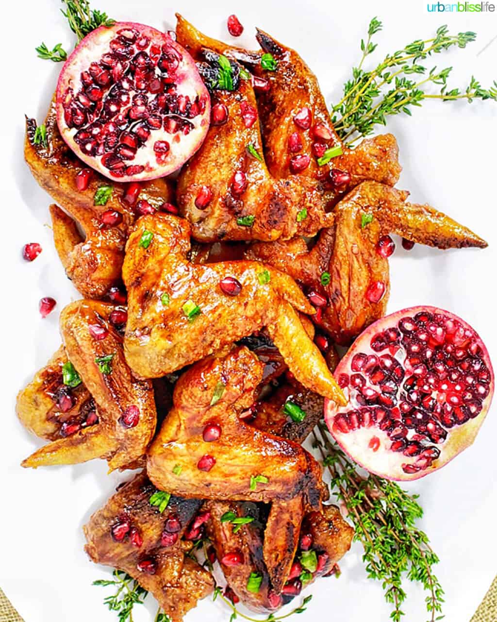 large platter of cranberry pomegranate sticky wings with a pomegranate halved on the side and herbs.