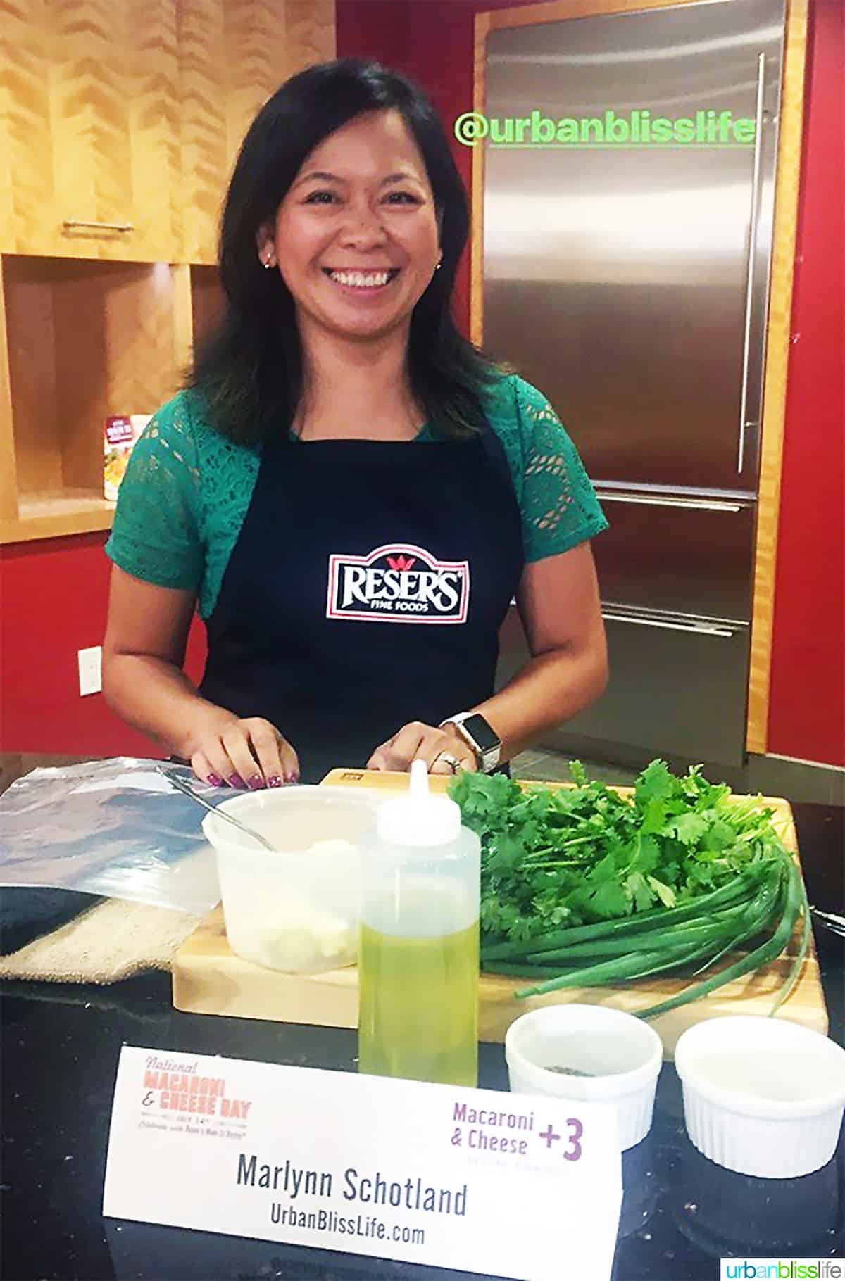 Marlynn Jayme Schotland in a Reser's apron with ingredients to make taco mac and cheese and a sign with her name and UrbanBlissLife.com. 