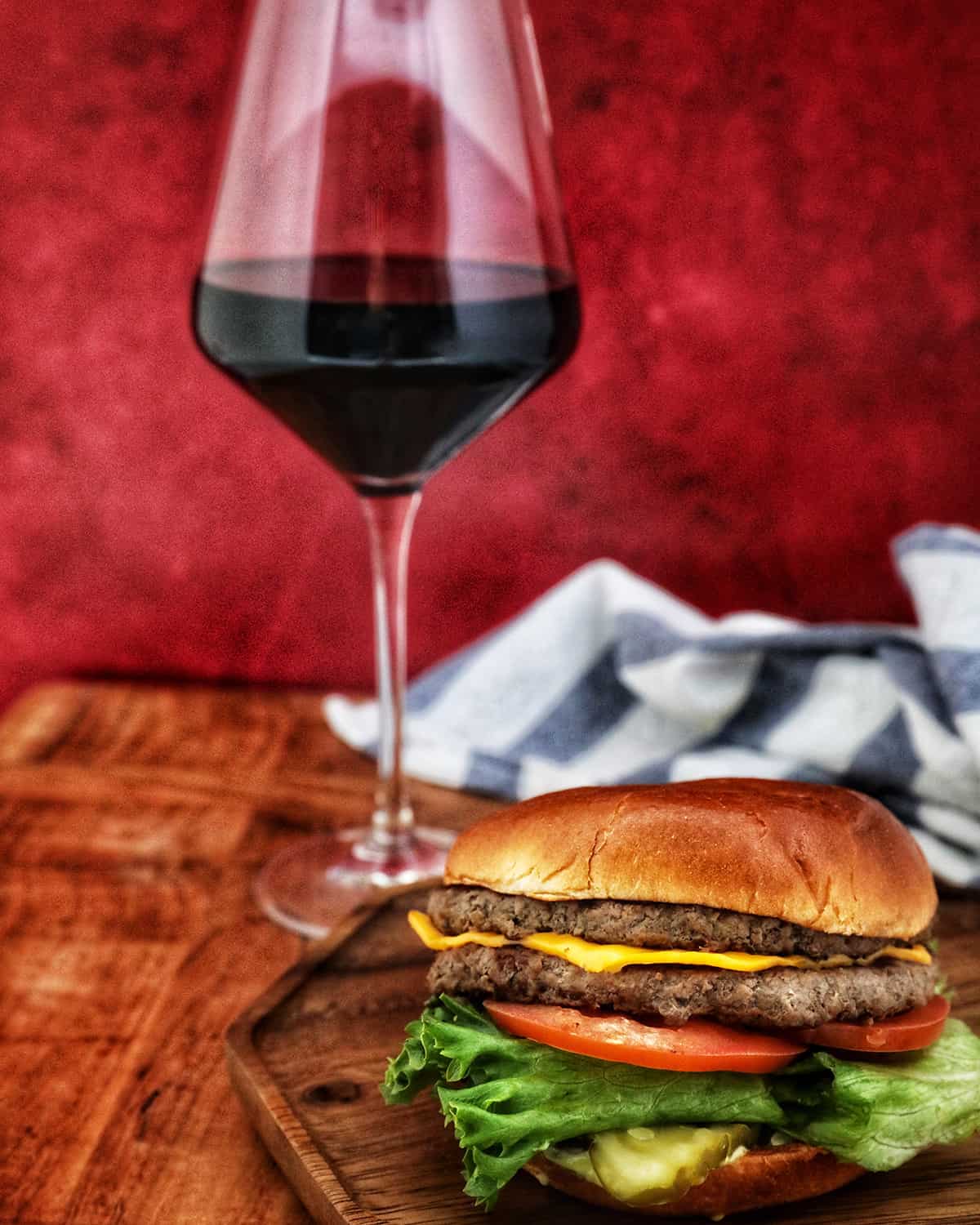 glass of Malbec behind a wooden tray with a loaded burger on top with red background.