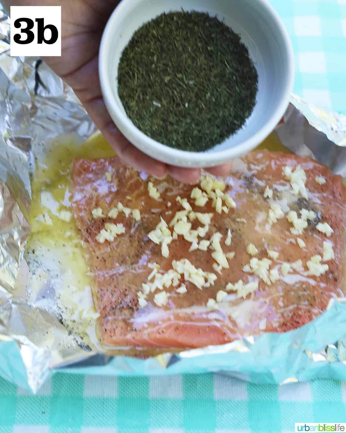 bowl of dill being poured onto salmon and garlic in foil on a checkered tablecloth.