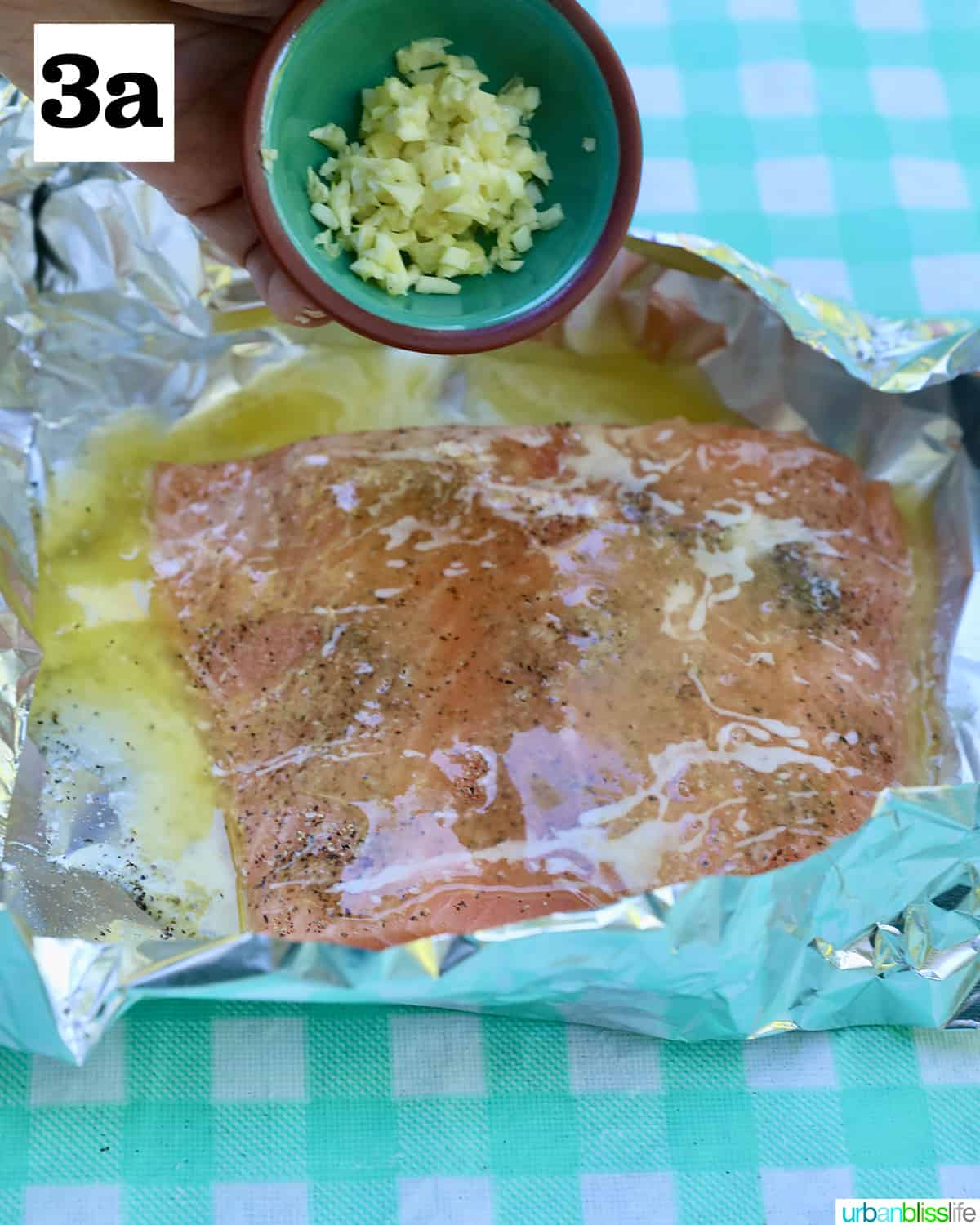 bowl of garlic added to seasoned salmon in foil on a checkered tablecloth.