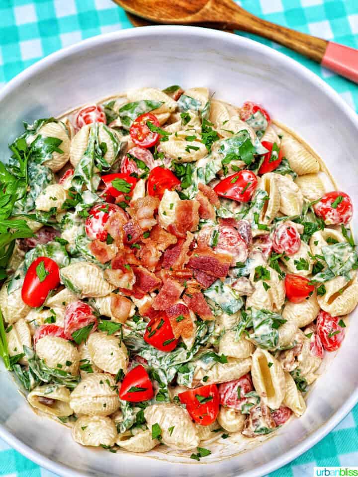 BLT pasta salad in a white bowl with wooden serving spoons overhead on an aqua checkered tablecloth.