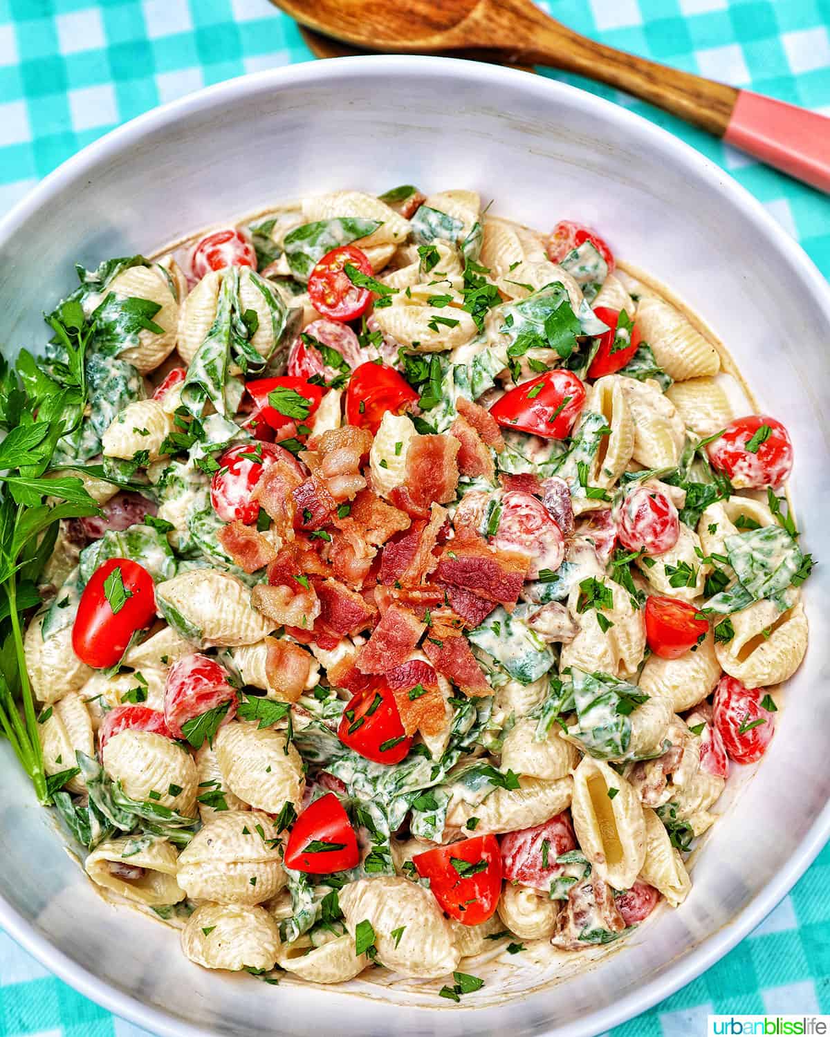 BLT pasta salad in a white bowl with wooden serving spoons overhead on an aqua checkered tablecloth.