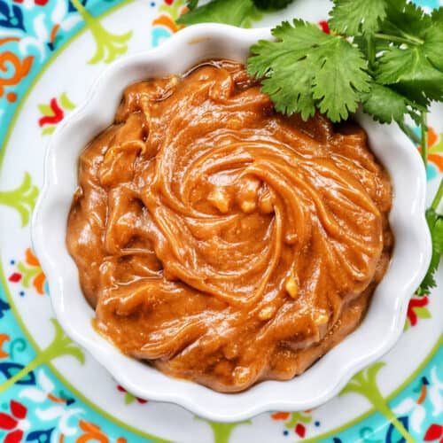 Thai peanut sauce in a white bowl with fresh cilantro leaves on a colorful patterned plate.