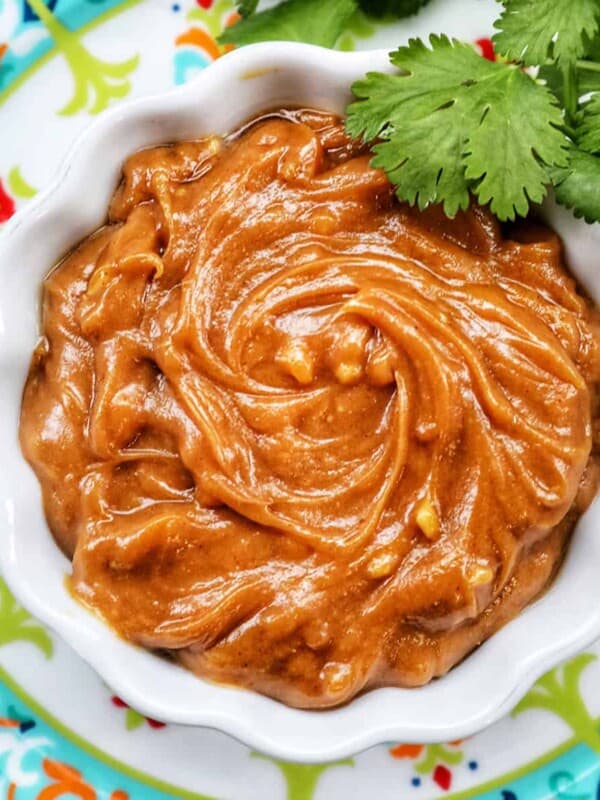 Thai peanut sauce in a white bowl with fresh cilantro leaves on a colorful patterned plate.
