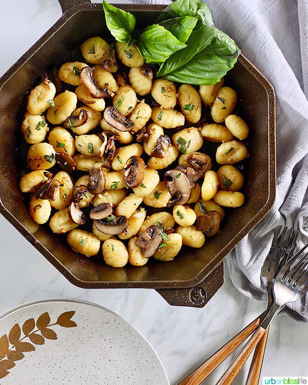 cast iron skillet with gnocchi, mushrooms, basil, plates and forks.