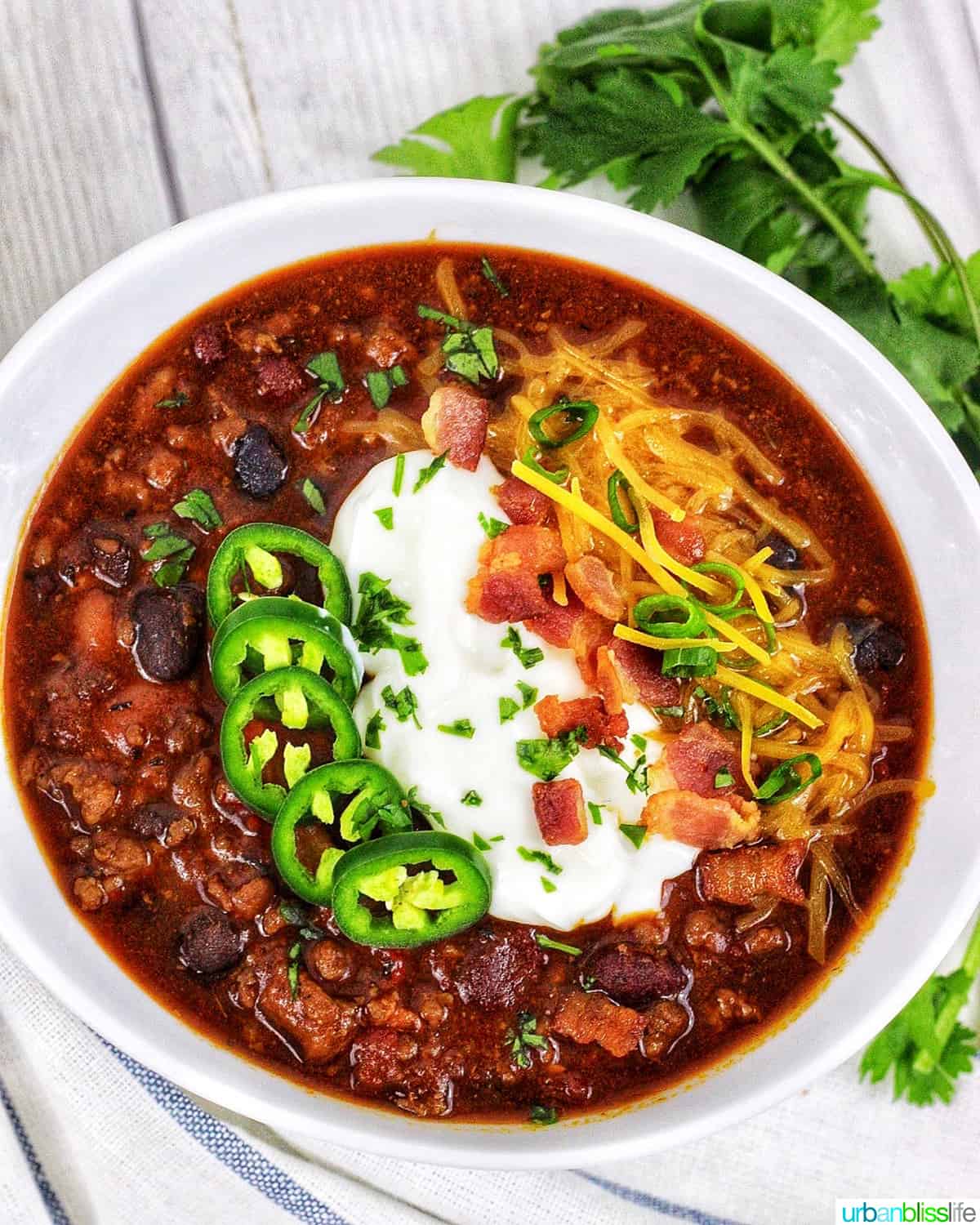 bowl of instant pot chili topped with jalapenos, sour cream, bacon, cheddar cheese, with green herbs on the side.