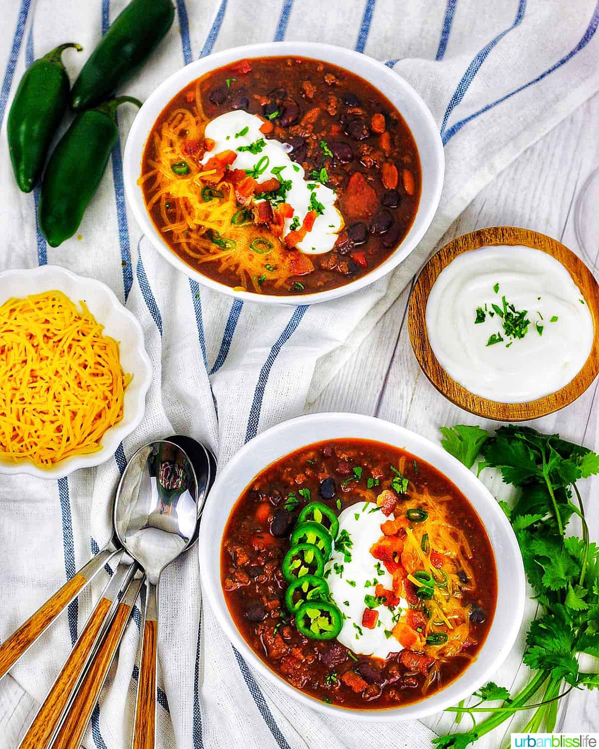two bowls of instant pot chili on white and blue napkin with sides of cheddar cheese, jalapenos, and sour cream.