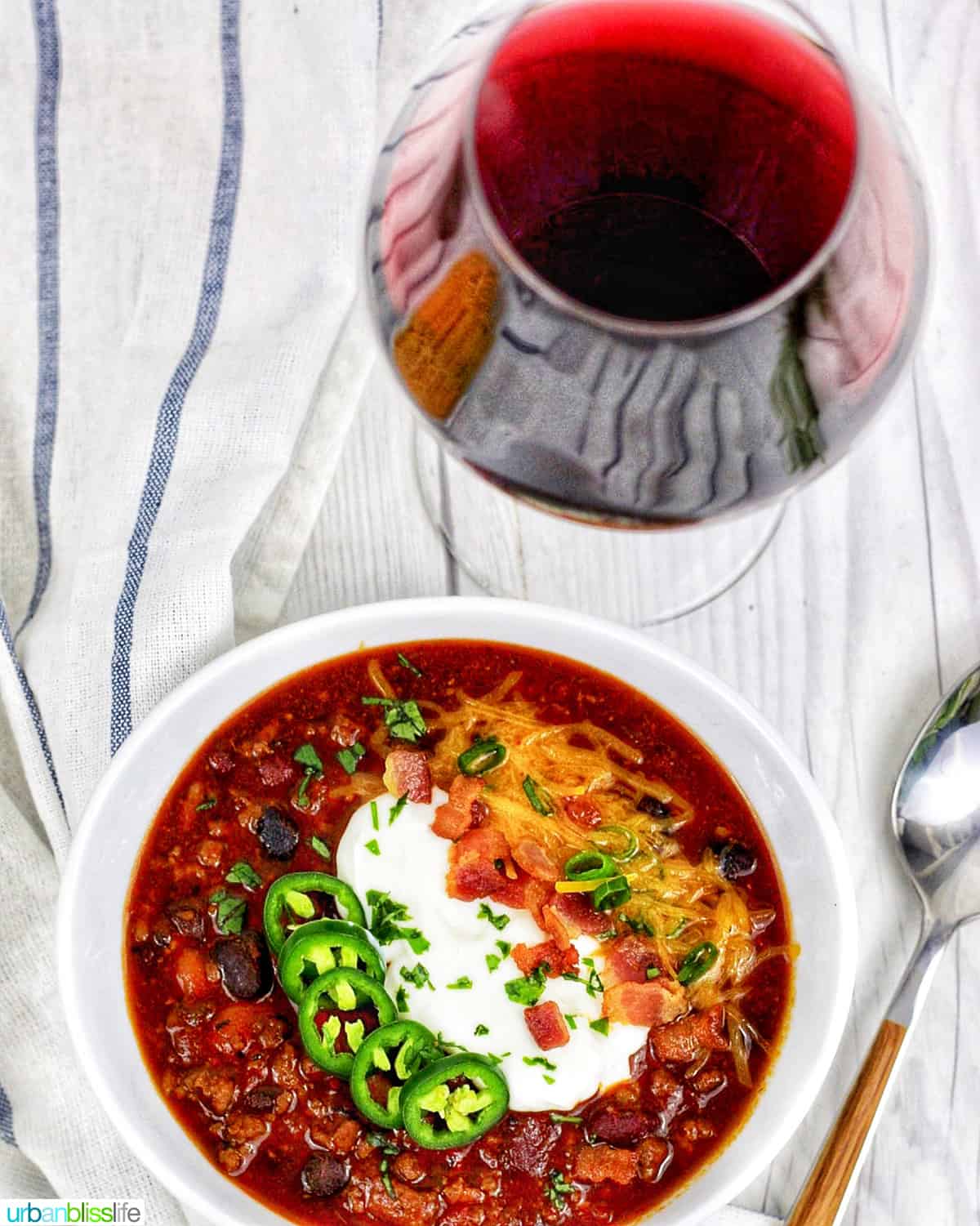 bowl of chili topped with jalapenos, sour cream, bacon, and cheese, with a glass of red wine on white table with spoon.