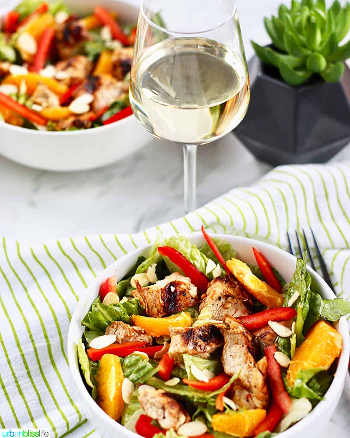 glass of white wine with two bowls of mandarin orange chicken salad with two forks and a green and white napkin.