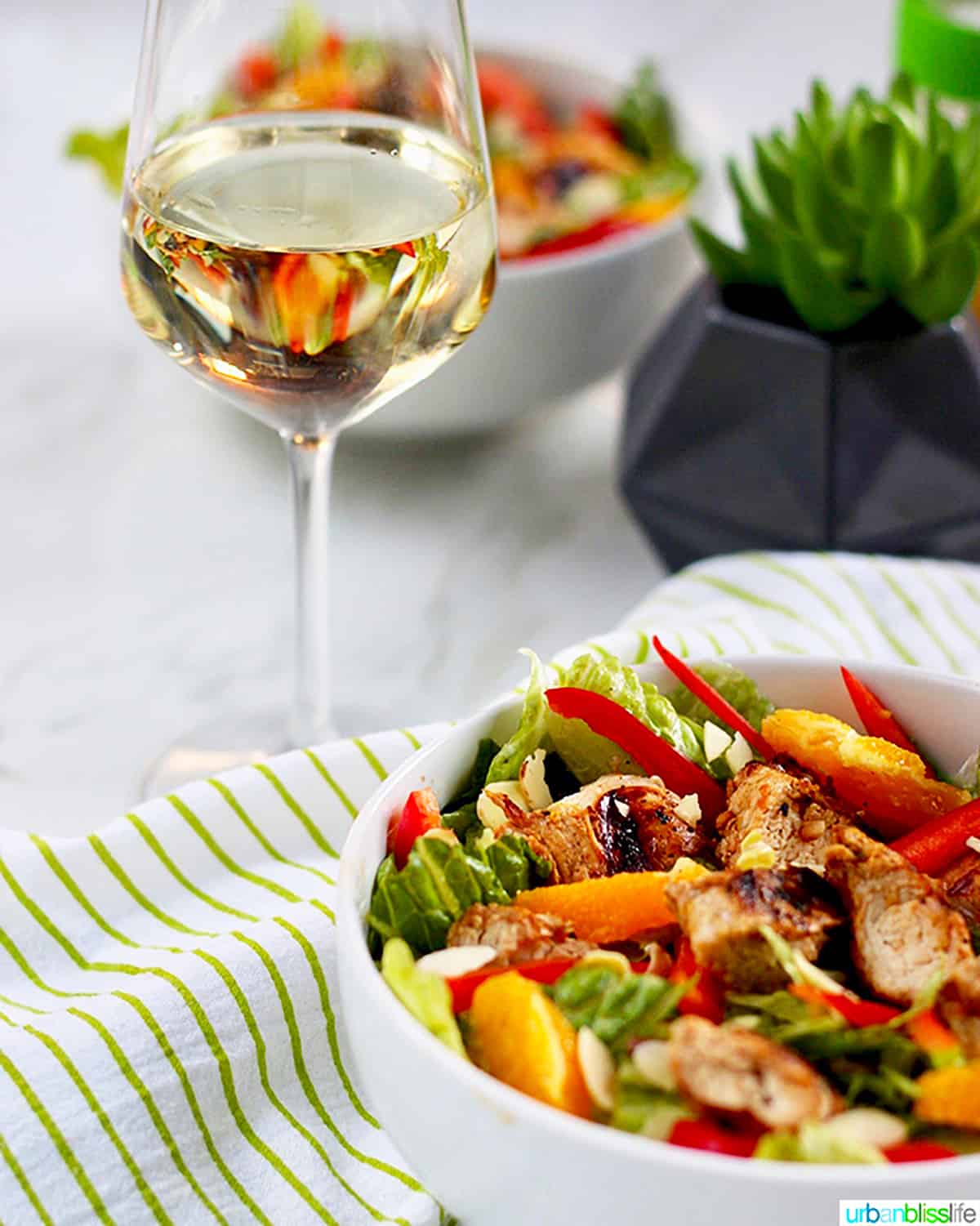 bowl of mandarin orange chicken salad with almonds on a bed of greens with glass of white wine.