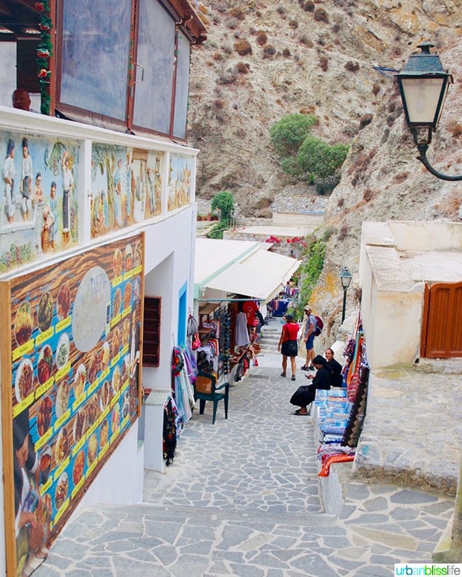 quaint side street with stone steps, restaurant with menu on the wall, and shop with clothes and accessories on Karpathos, Greece.