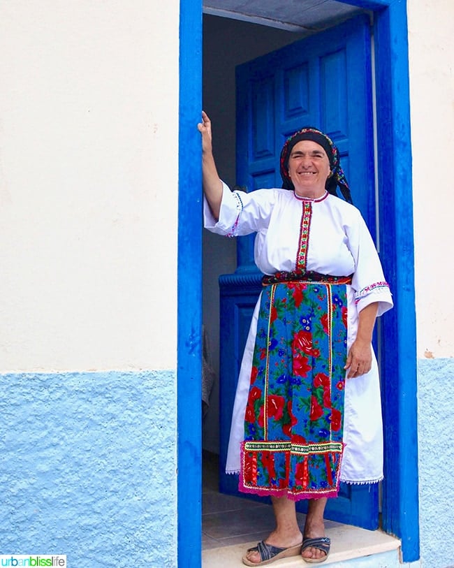 Karpathian woman dressed in traditional white and floral dress with colorful floral scarf standing in doorway on Karpathos, Greece.