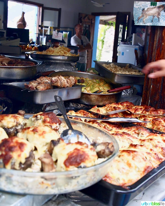 food stall on Karpathos, Greece with trays full of Greek food and shop owner in the background.