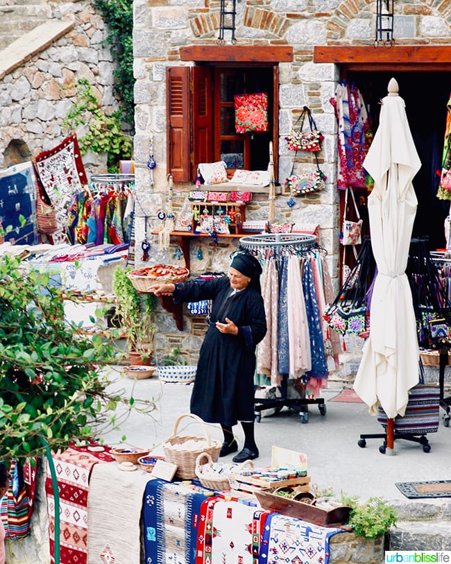 old woman dressed in all black standing in a market surrounded by colorful rugs, clothes, scarves, and bags on Karpathos, Greece.