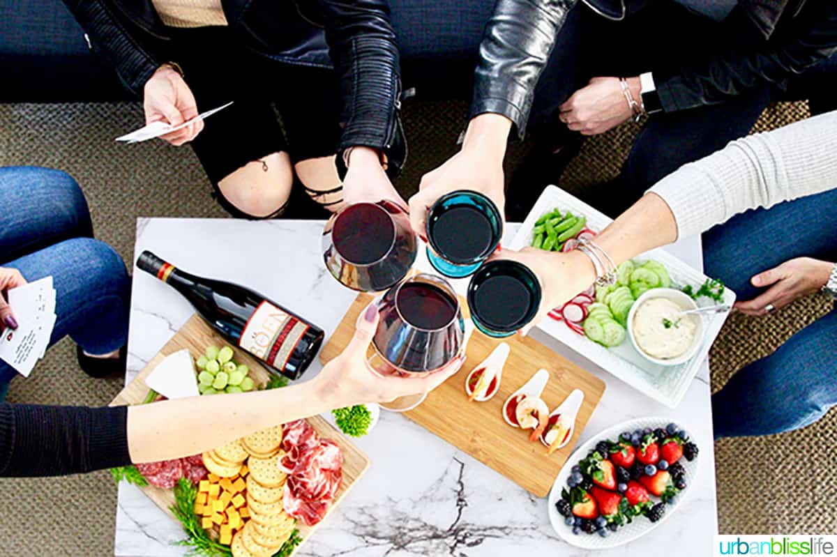 four women leaning in to cheers with glasses of wine over a table with charcuterie boards and holding game cards.