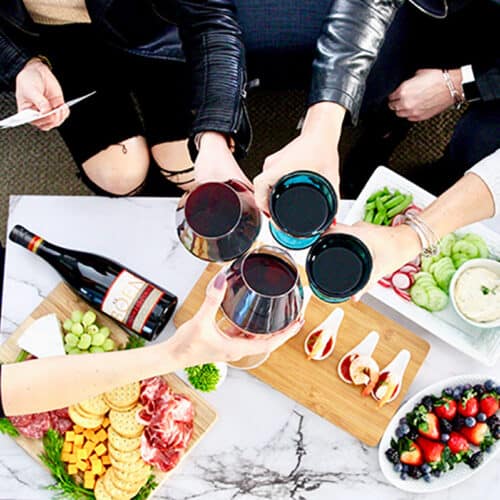 four women leaning in to cheers with glasses of wine over a table with charcuterie boards and holding game cards.
