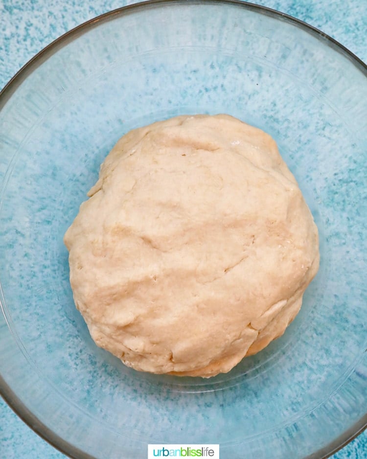 raw pizza dough ball in a clear glass bowl on top of a blue table.