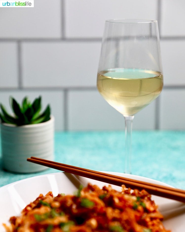 glass of white wine with noodles in a white bowl and chopsticks on the side.