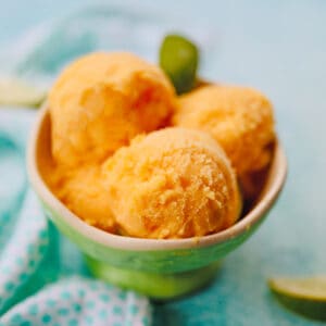 bowl of scoops of pineapple mango sorbet on a bright blue background with napkin and limes on the side.