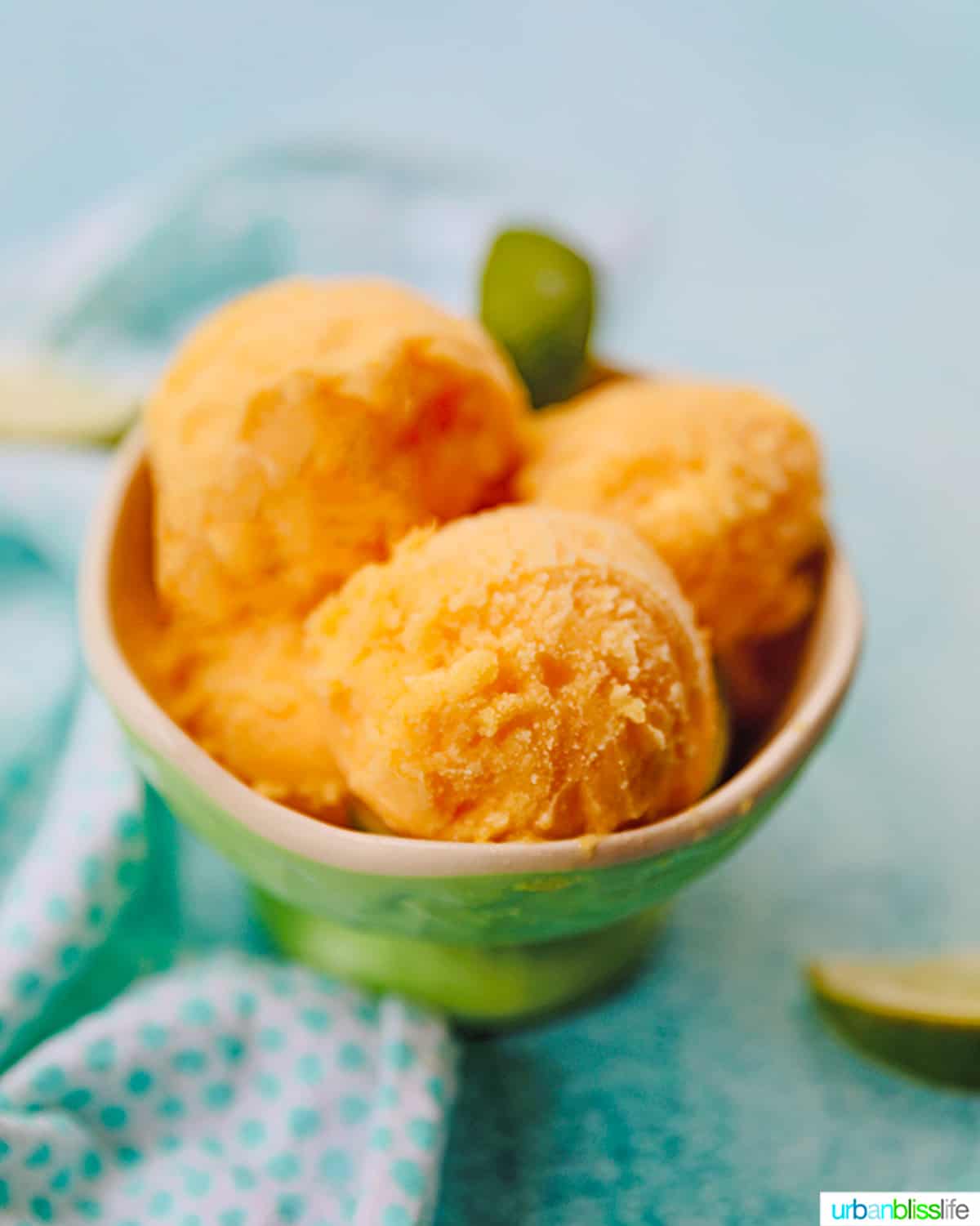 bowl of scoops of pineapple mango sorbet on a bright blue background with napkin and limes on the side.