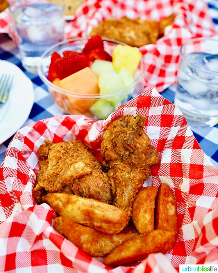 summer outdoor picnic with fried chicken, jo jos, cookies, fruit, on a blue and white checkered tablecloth