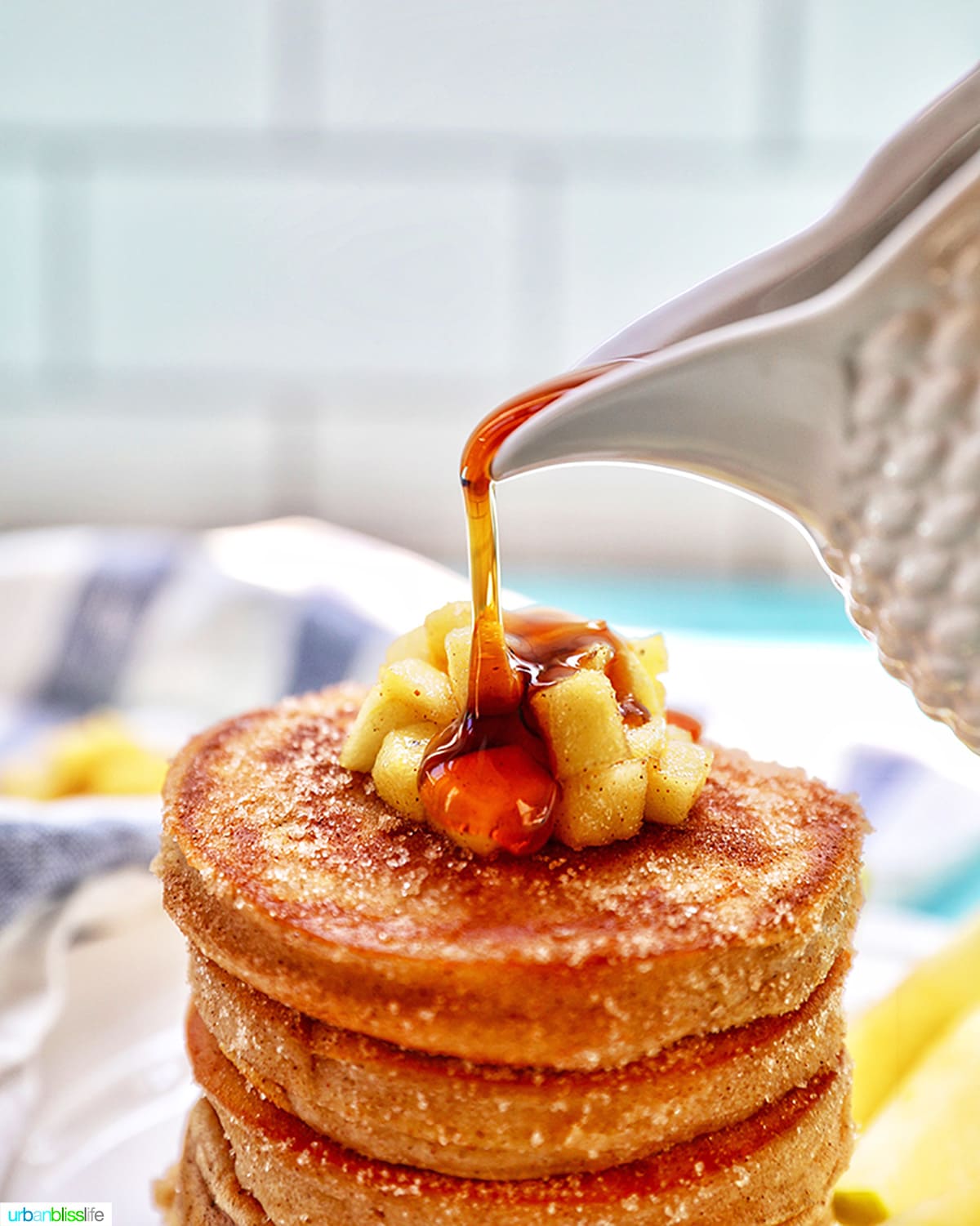 pouring maple syrup over a stack of pancakes with chopped apples on top.