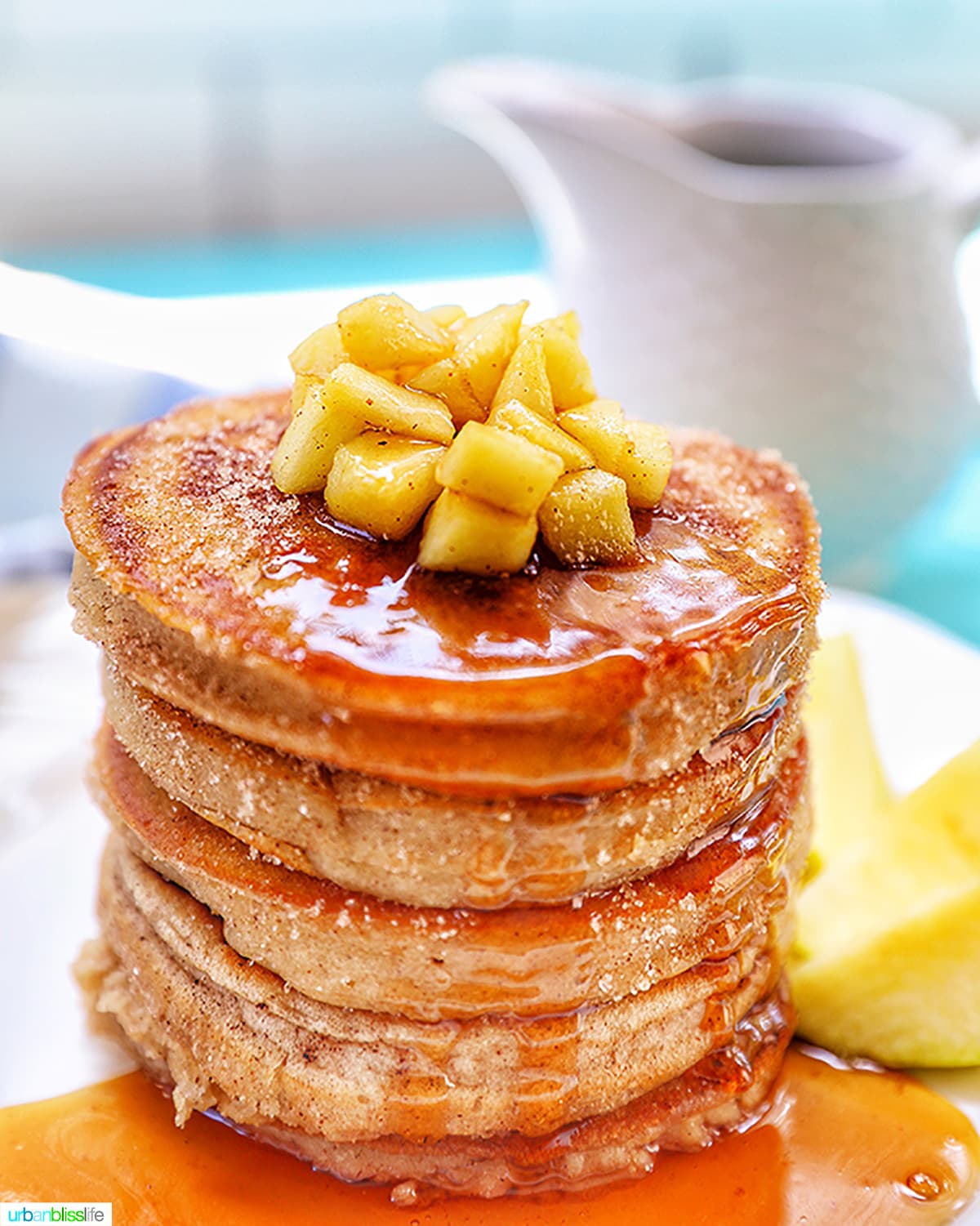 stack of apple cinnamon pancakes with syrup and chopped apples on top, white jar of syrup in the background.