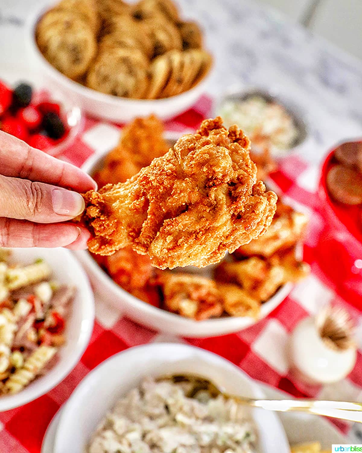 holding a fried chicken wing up close with tailgating food in background