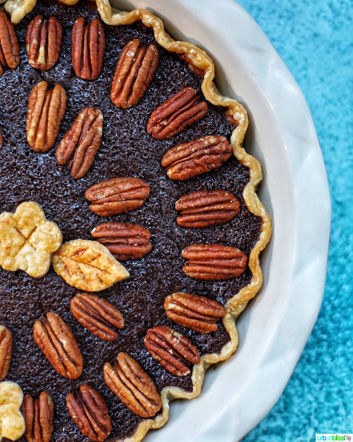 right half of Chocolate Bourbon Pecan Pie with pie crust cutouts in leaf and flower shapes with pecan halves as pie decoration in a white pie pan on blue table.