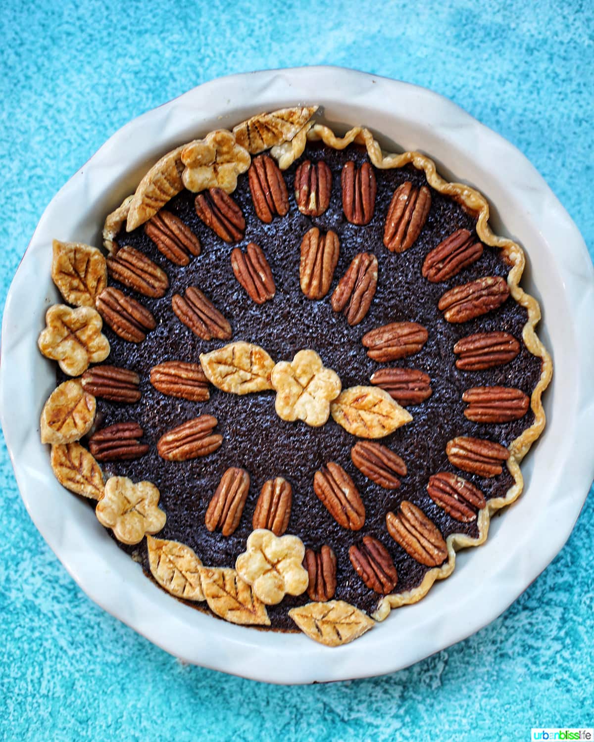 Chocolate Bourbon Pecan Pie with pie crust cutouts in leaf and flower shapes with pecan halves as pie decoration in a white pie pan on blue table.