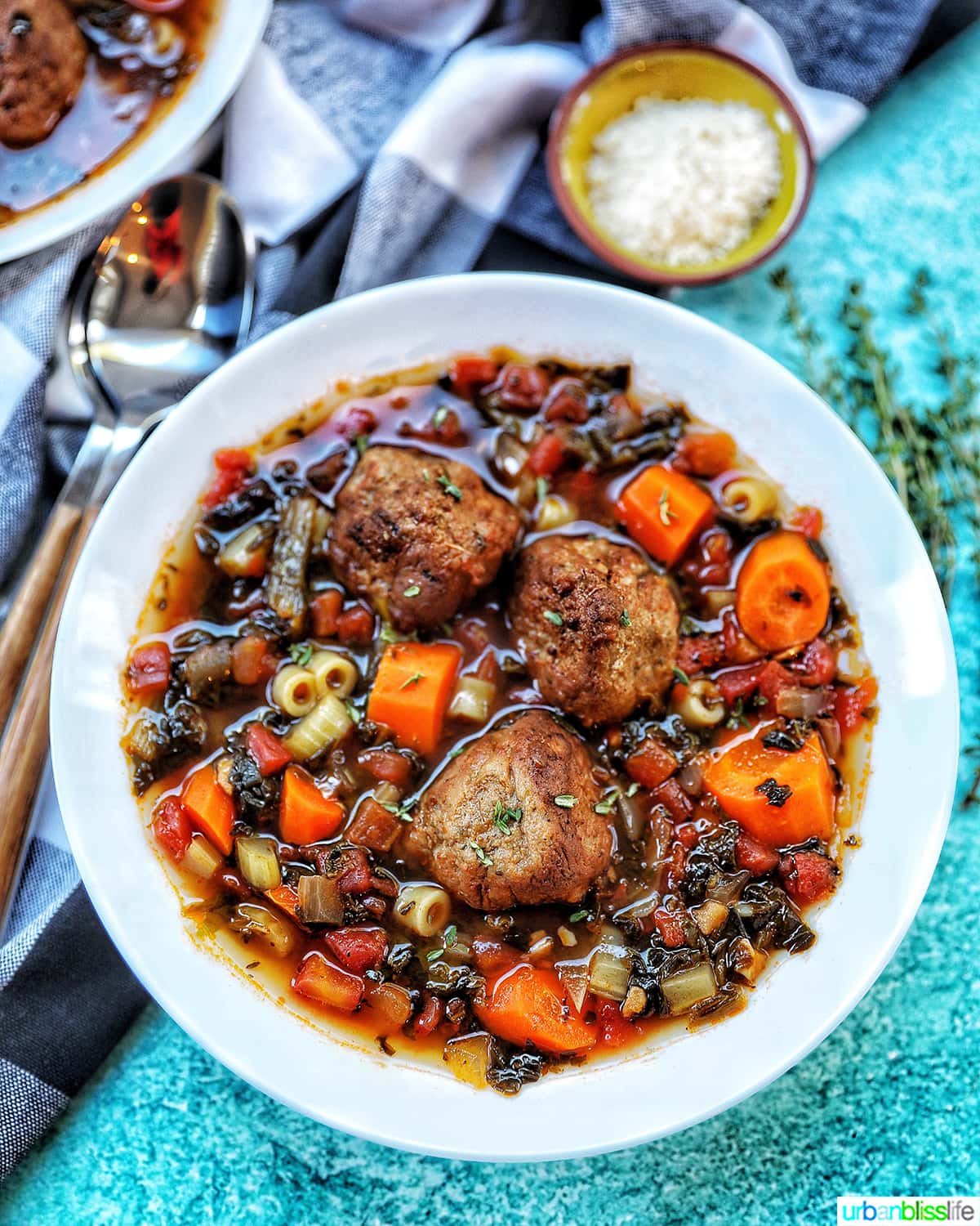 large white bowl of meatball soup with carrots, pasta, and a side of parmesan cheese and spoons.