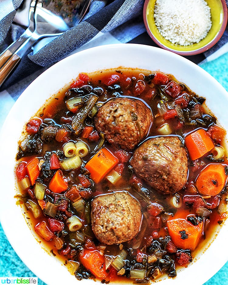 large white bowl of meatball soup with carrots, pasta, and a side of parmesan cheese and spoons.